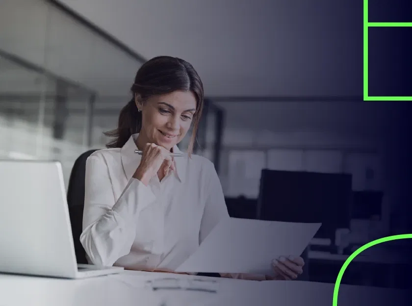 Mulher em uma mesa de escritório observando um papel, com uma caneta na mão.