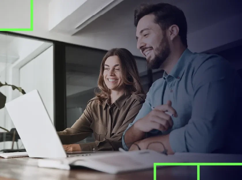 Foto de um homem e uma mulher em um ambiente corporativo, sorrindo e olhando para o computador - Como criar um banco digital